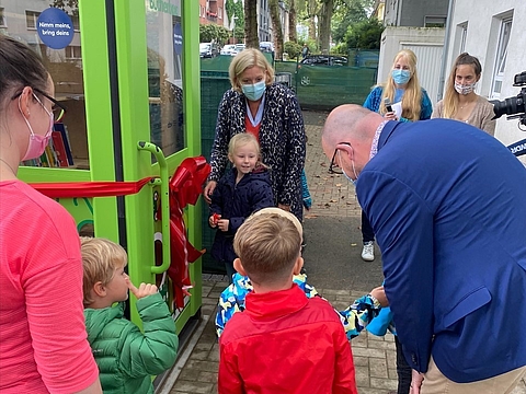 Kinder und Erwachsene lösen gemeinsam die große Schleife an der Kinder-Bücherbox, um sie zu öffnen