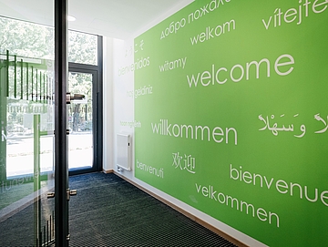 Flur eines Kindergartens mit Glastür, die den Blick nach außen freigibt. Auf einer grünen Wand steht das Wort "Willkommen" in verschiedenen Sprachen